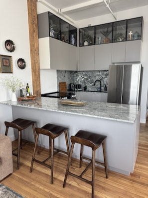 A modern kitchen with a central island, bar stools, a stainless steel refrigerator, and a marble backsplash. - Room 307, Ilium Luxury 2 Bd. (Troy)