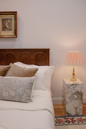 Een net opgemaakt bed met kussens, een houten hoofdbord, een bedlampje op een marmeren nachtkastje en een ingelijste foto aan de muur.