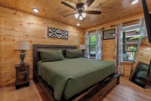 Een slaapkamer met houten lambrisering, een bed, bedlampen, een plafondventilator en een raam met gordijnen.