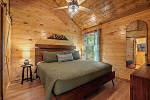 Een slaapkamer in een houten hutje, met een bed, nachtkastje, lamp, plafondventilator en een raam met gordijnen.