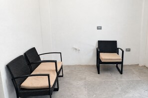 Deux fauteuils en osier noir, dotés de coussins beige, dont l’un a un dossier et l’autre un coussin d’assise, sont placés contre un mur blanc, au-dessus d’un sol carrelé.