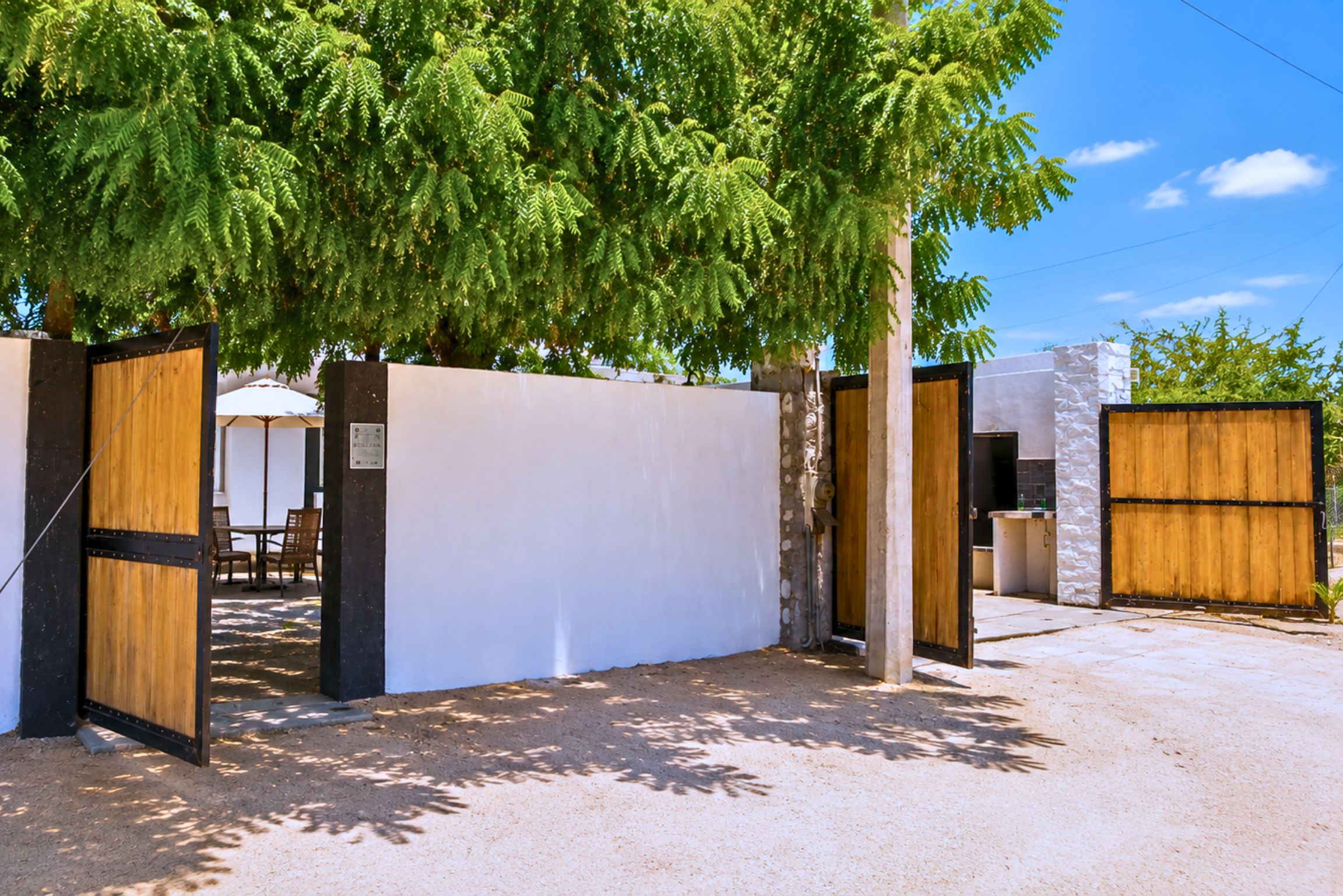 A gated entrance with a white wall and a wooden door, leading to a covered outdoor seating area with tables and chairs.