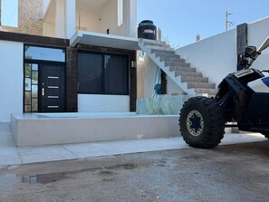 Un bâtiment moderne avec un escalier menant à une porte, un véhicule garé devant et un réservoir d’eau sur le toit.