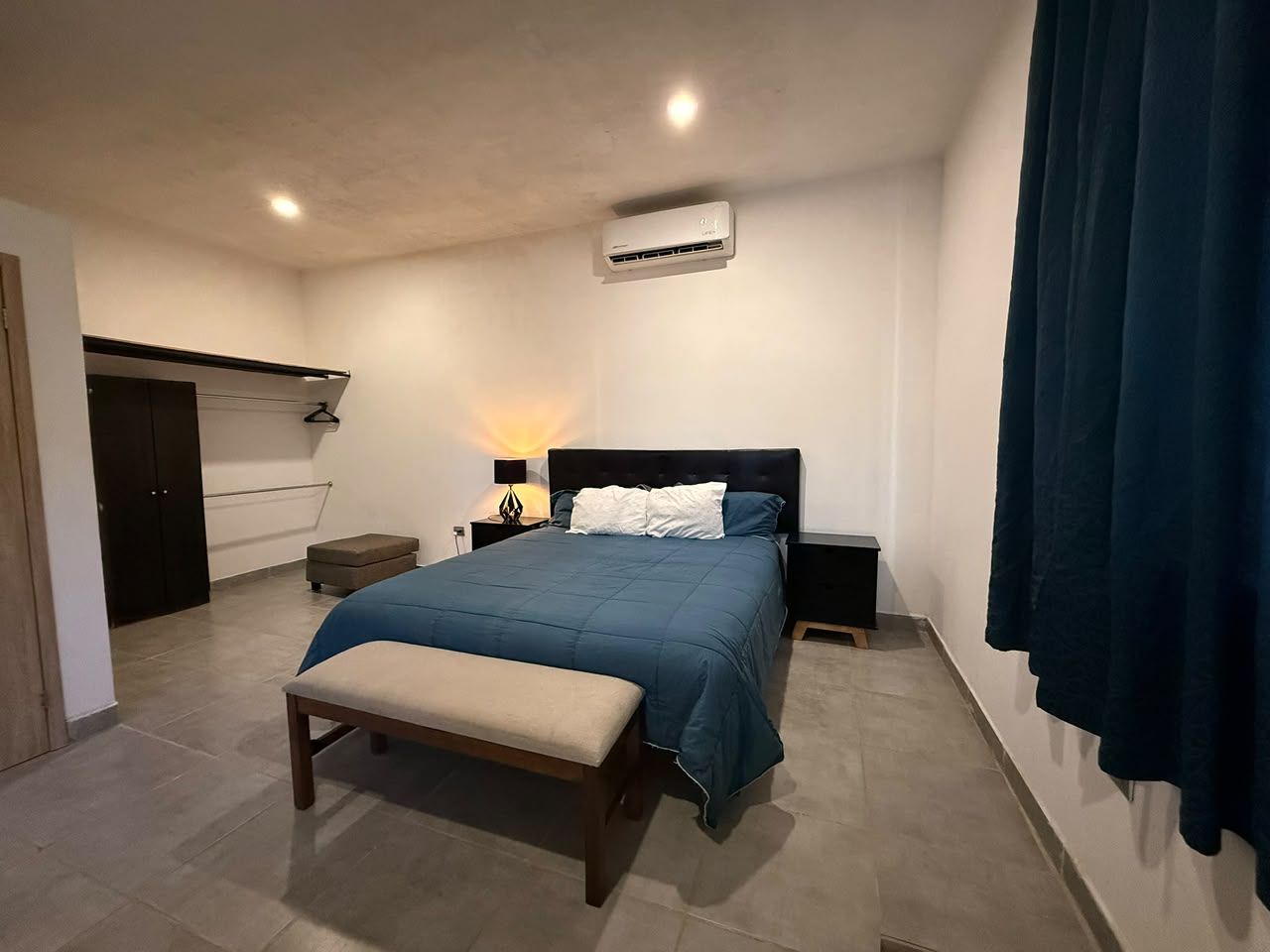 Quarto com cama, mesas de cabeceira, um banco, ar condicionado e guarda-roupa.