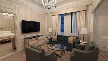 A hotel room with a bed, a TV, a sofa, two armchairs, a glass coffee table, a chandelier, and a window with curtains.