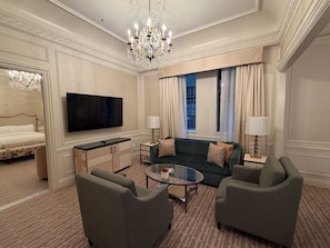 Een hotelkamer met een bed, een televisie, een bank, twee fauteuils, een glazen salontafel, een kroonluchter en een raam met gordijnen.