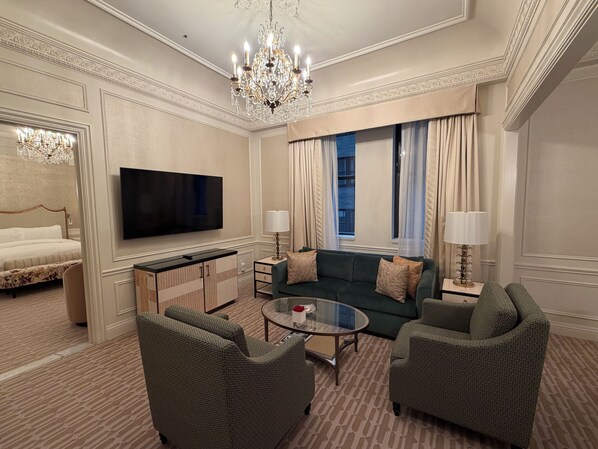 Une chambre d’hôtel comprenant un lit, une télévision, un canapé, deux fauteuils, une table basse en verre, un lustre et une fenêtre avec des rideaux.