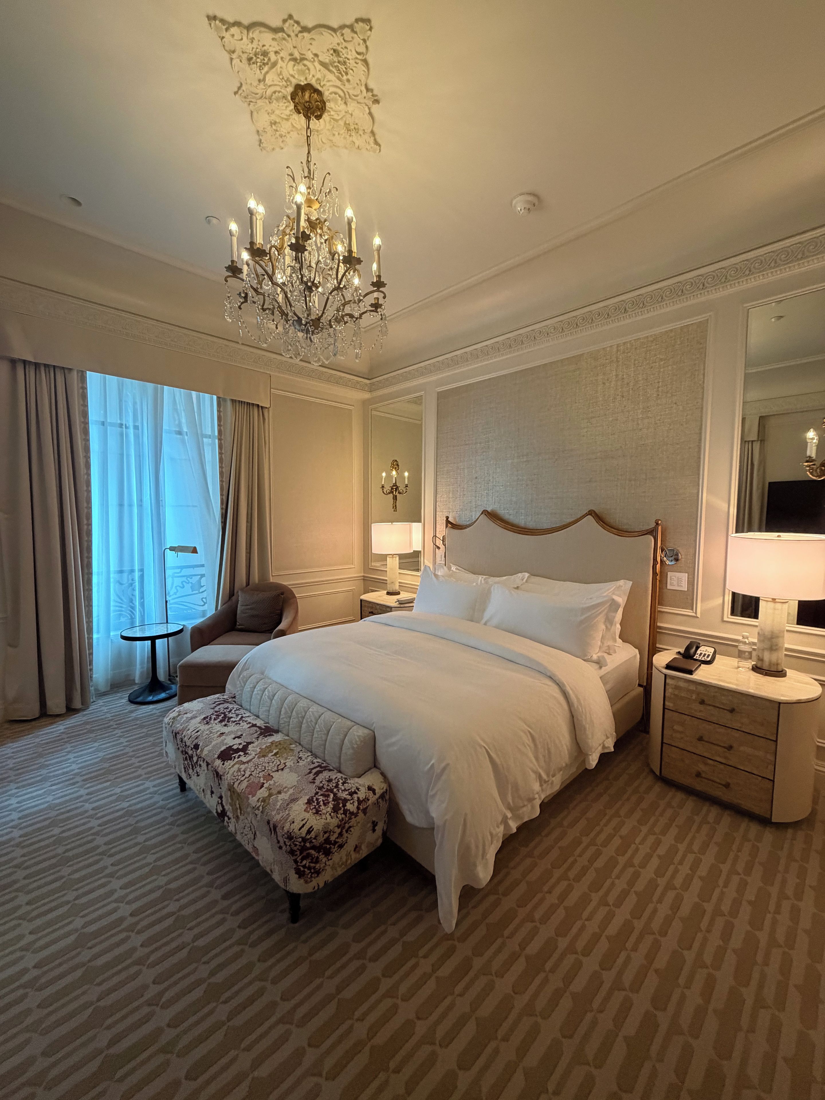 A hotel room with a large bed, a sitting area with a chair, a small table, and a nightstand with a lamp.