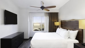 Quarto de hotel com cama, TV de tela plana, ventilador de teto, janela com cortinas e mesa de cabeceira com abajur.