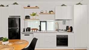 A modern kitchen with white cabinetry, a black refrigerator, a built-in oven, and a dining area with a wooden table and chairs. - 10 Hobson - Coastal Retreat (Queenscliff)