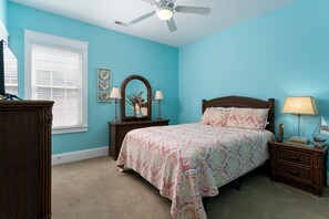 Ein Schlafzimmer mit einem Bett, einem Schminktisch, einem Nachttisch, einem Deckenventilator und einem Fenster mit Jalousien.
