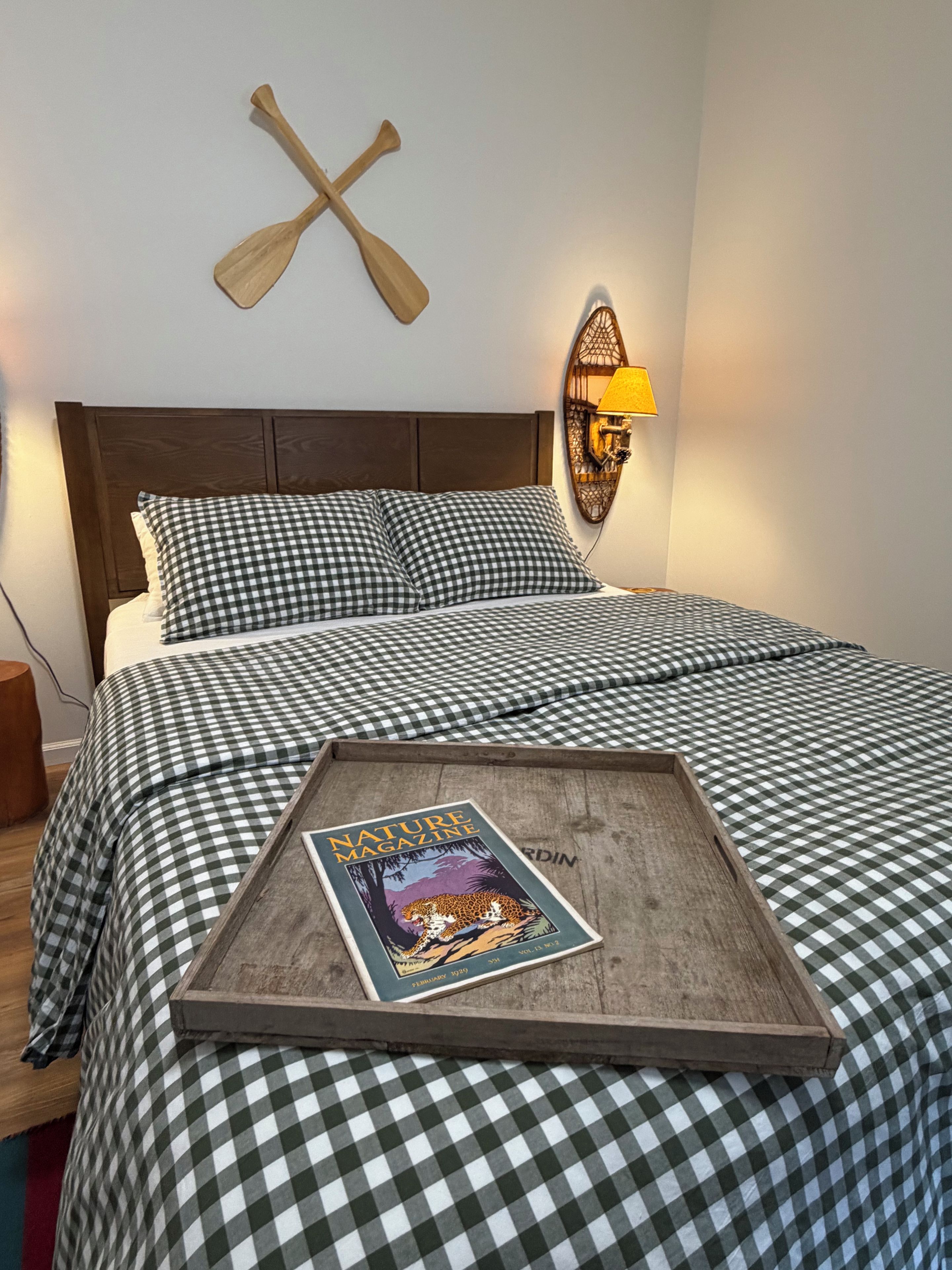 A neatly made bed with checkered bedding, a magazine on the bed, and a wall-mounted lamp.
