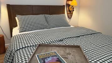 A neatly made bed with checkered bedding, a magazine on the bed, and a wall-mounted lamp.