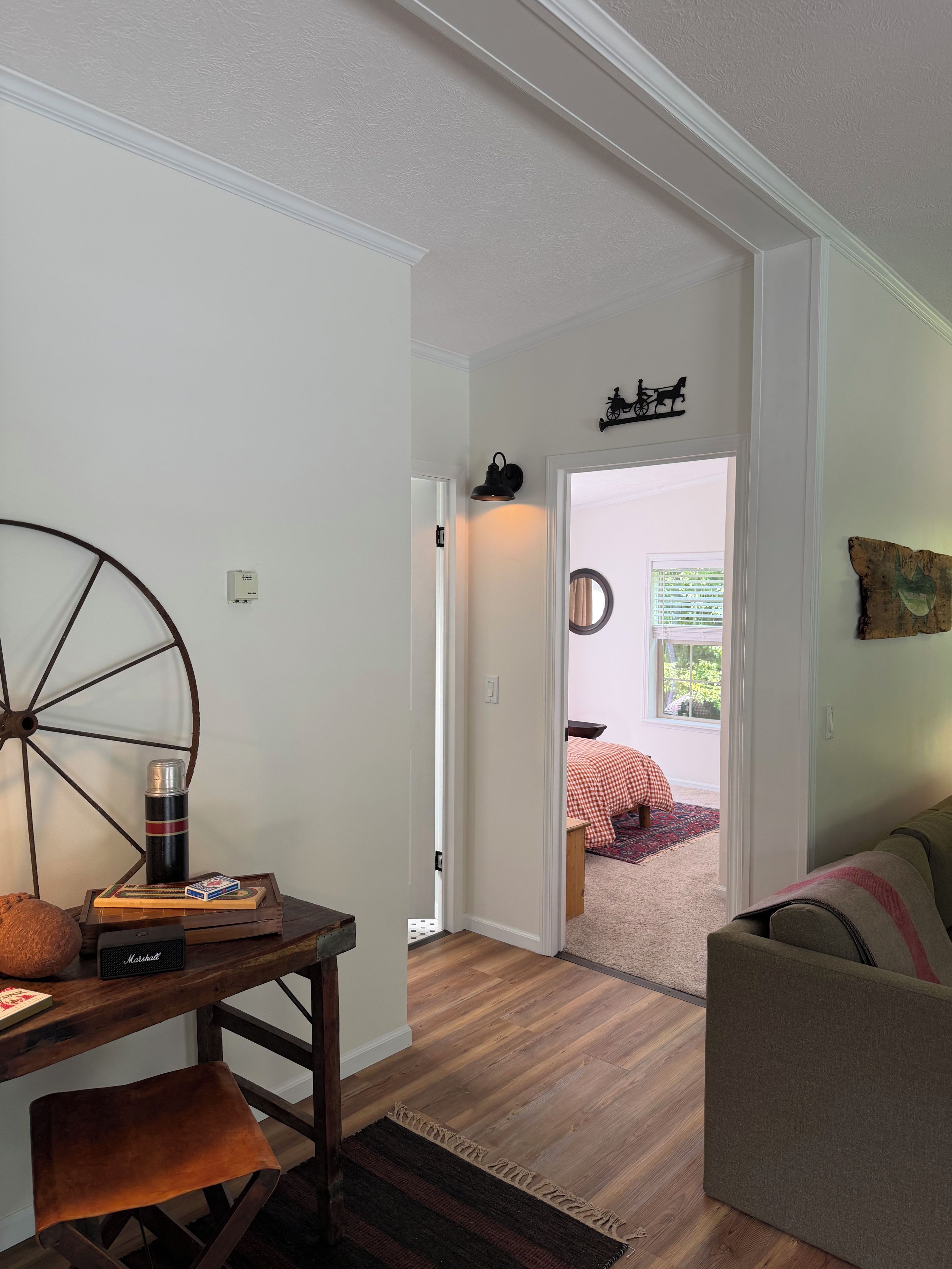 Quarto com mesa de madeira, uma cadeira, radiador, cama com colcha xadrez e janela com vista para o verde.