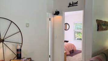 Quarto com mesa de madeira, uma cadeira, radiador, cama com colcha xadrez e janela com vista para o verde.