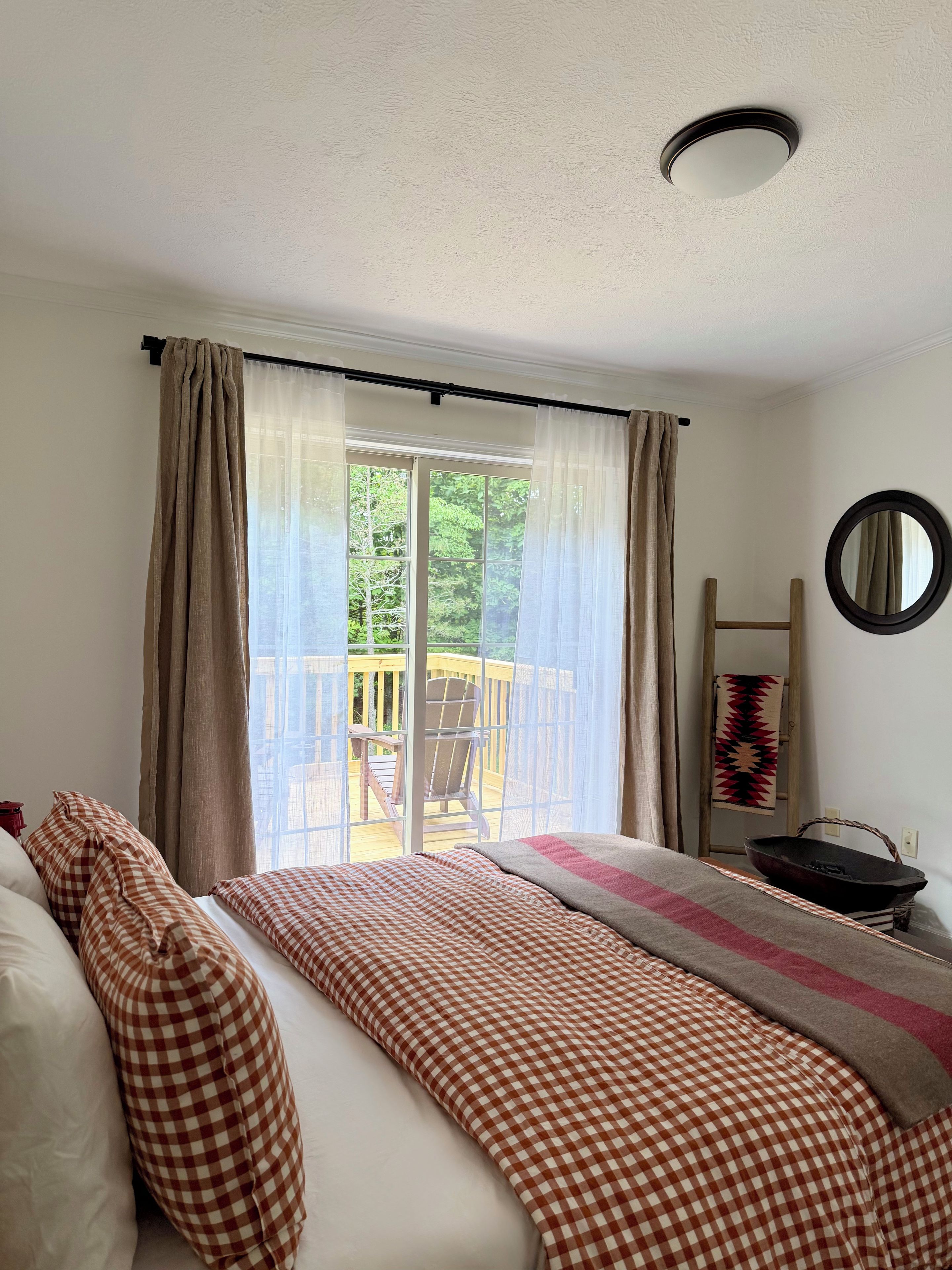 A bedroom with a bed, checkered bedding, a window with curtains, a view of a deck, and a round mirror on the wall.