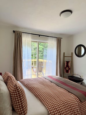 A bedroom with a bed, checkered bedding, a window with curtains, a view of a deck, and a round mirror on the wall.