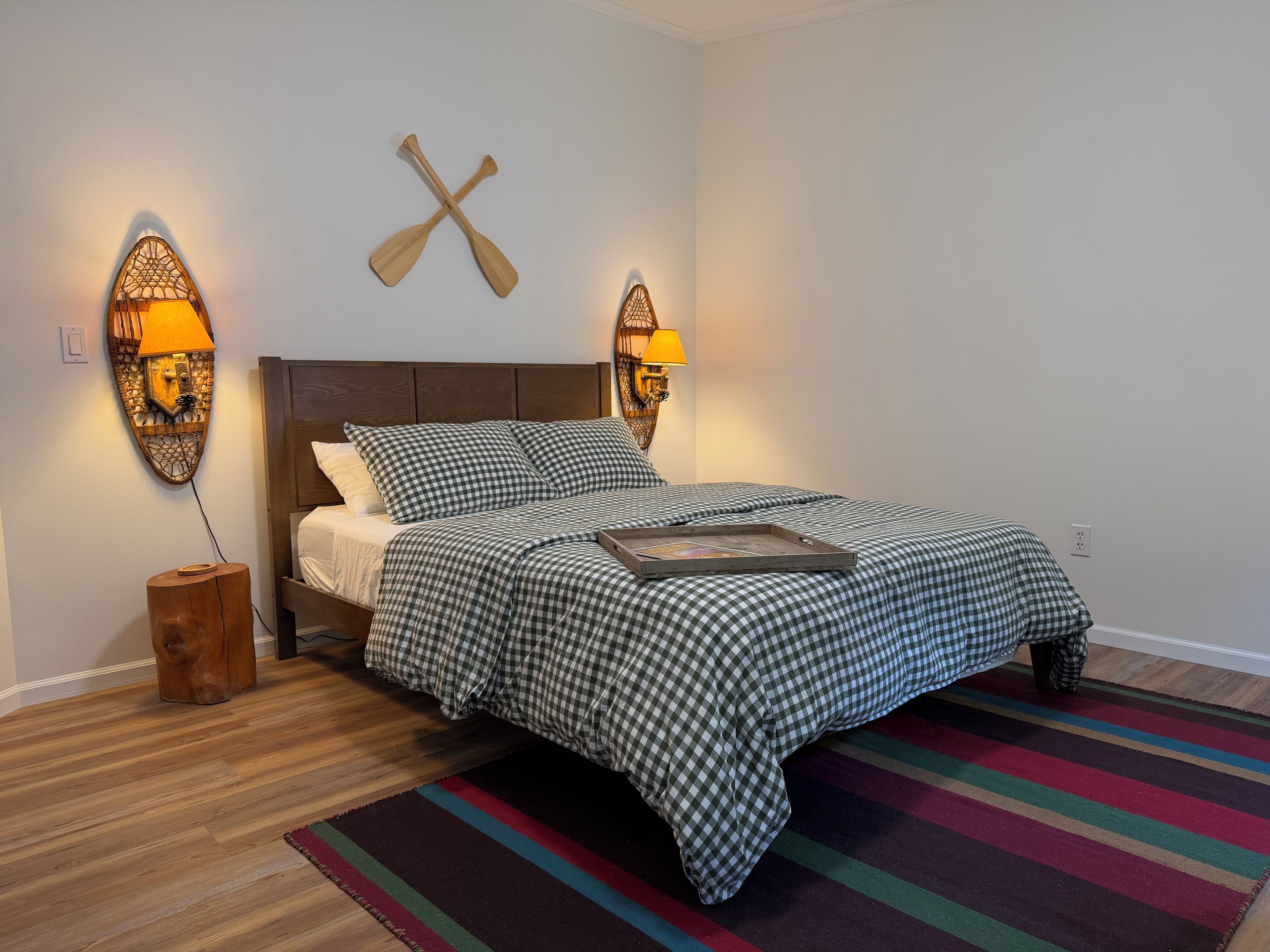 Cama bem arrumada com colcha xadrez, mesa de cabeceira de madeira com abajur e um remo de madeira fixado na parede.