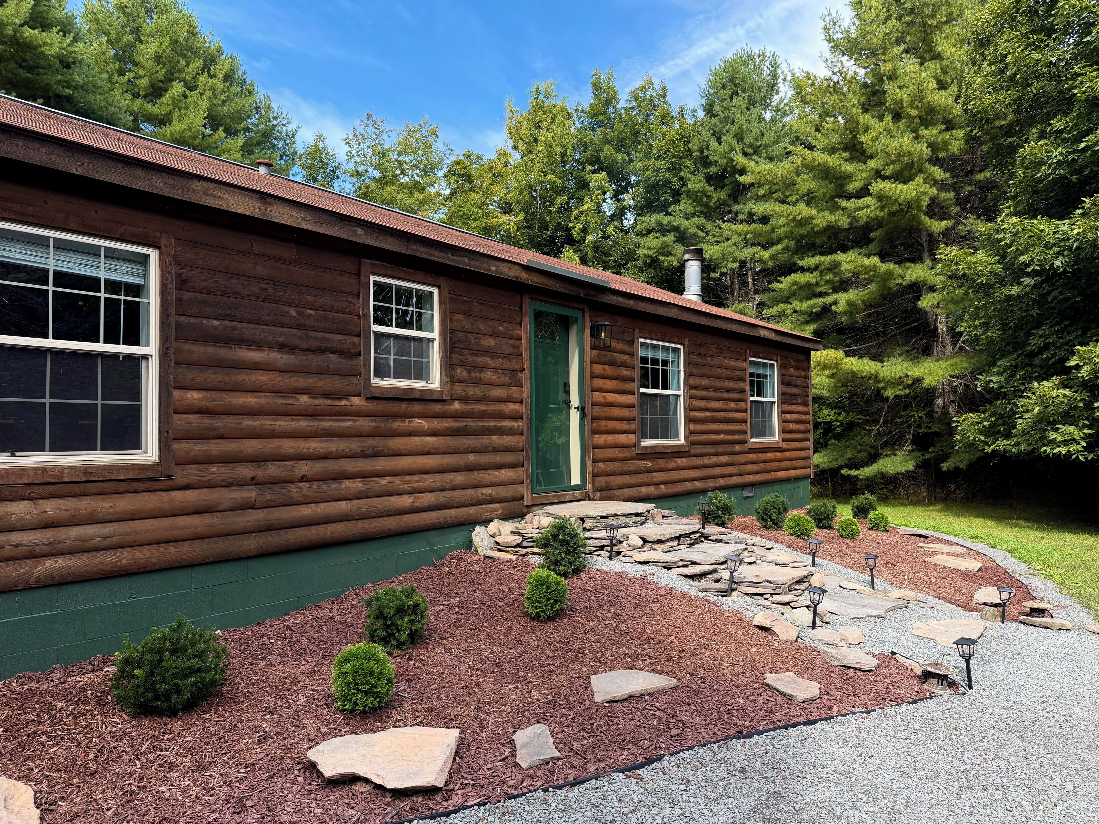 A log cabin with a green door, two windows, and a stone pathway leading to the entrance.