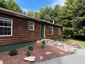 A log cabin with a green door, two windows, and a stone pathway leading to the entrance.