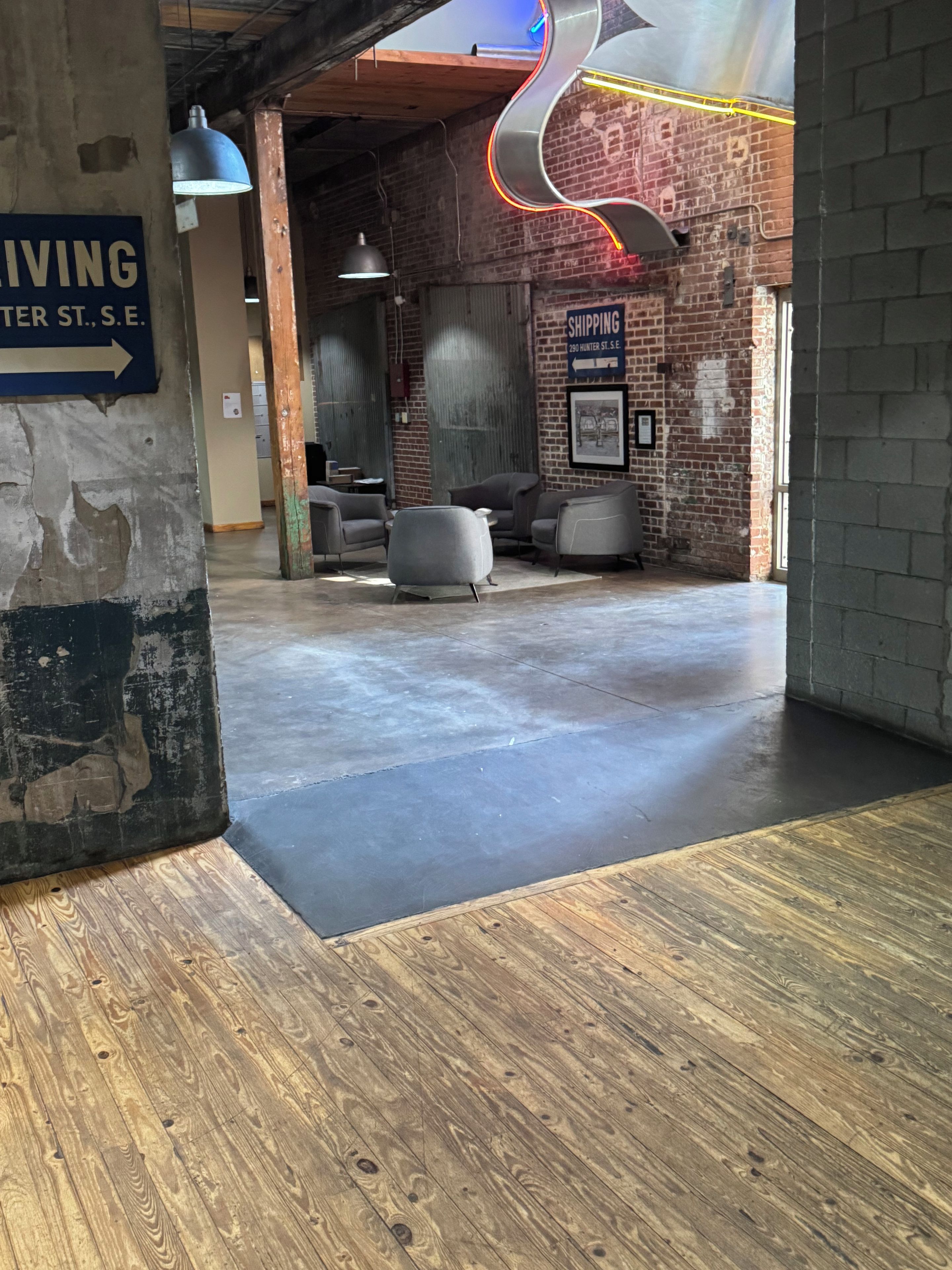 Un interior amplio con piso de concreto, detalles de madera y una pared de ladrillo. Hay zonas de estar con sillas modernas y un letrero de neón.