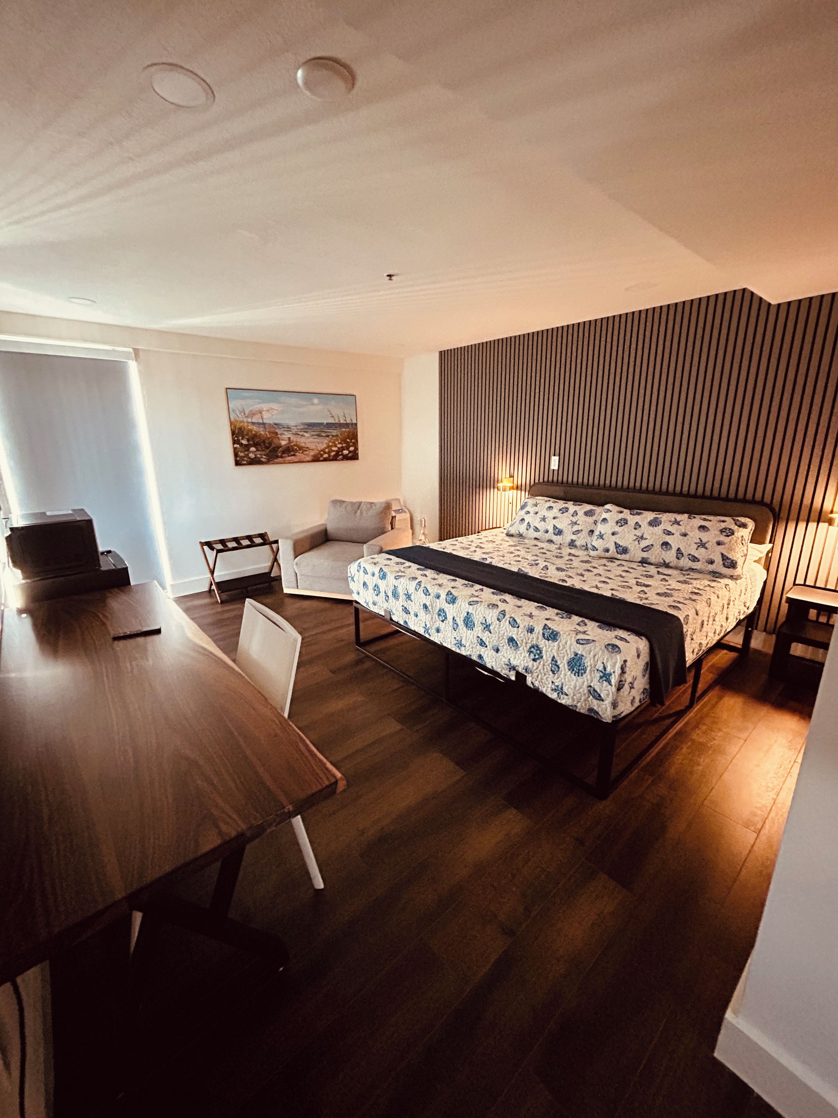 A bedroom with a bed, a desk, a chair, a bedside table, and a wall-mounted lamp.