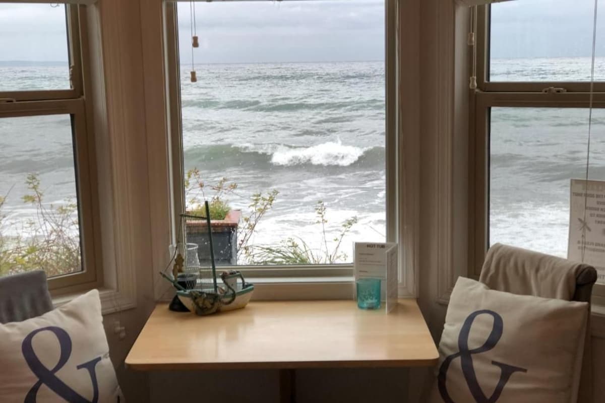 Une chambre avec vue sur l’océan, grâce à de grandes fenêtres, une petite table sur laquelle se trouve un récipient en verre, et deux coussins ornés d’un monogramme.
