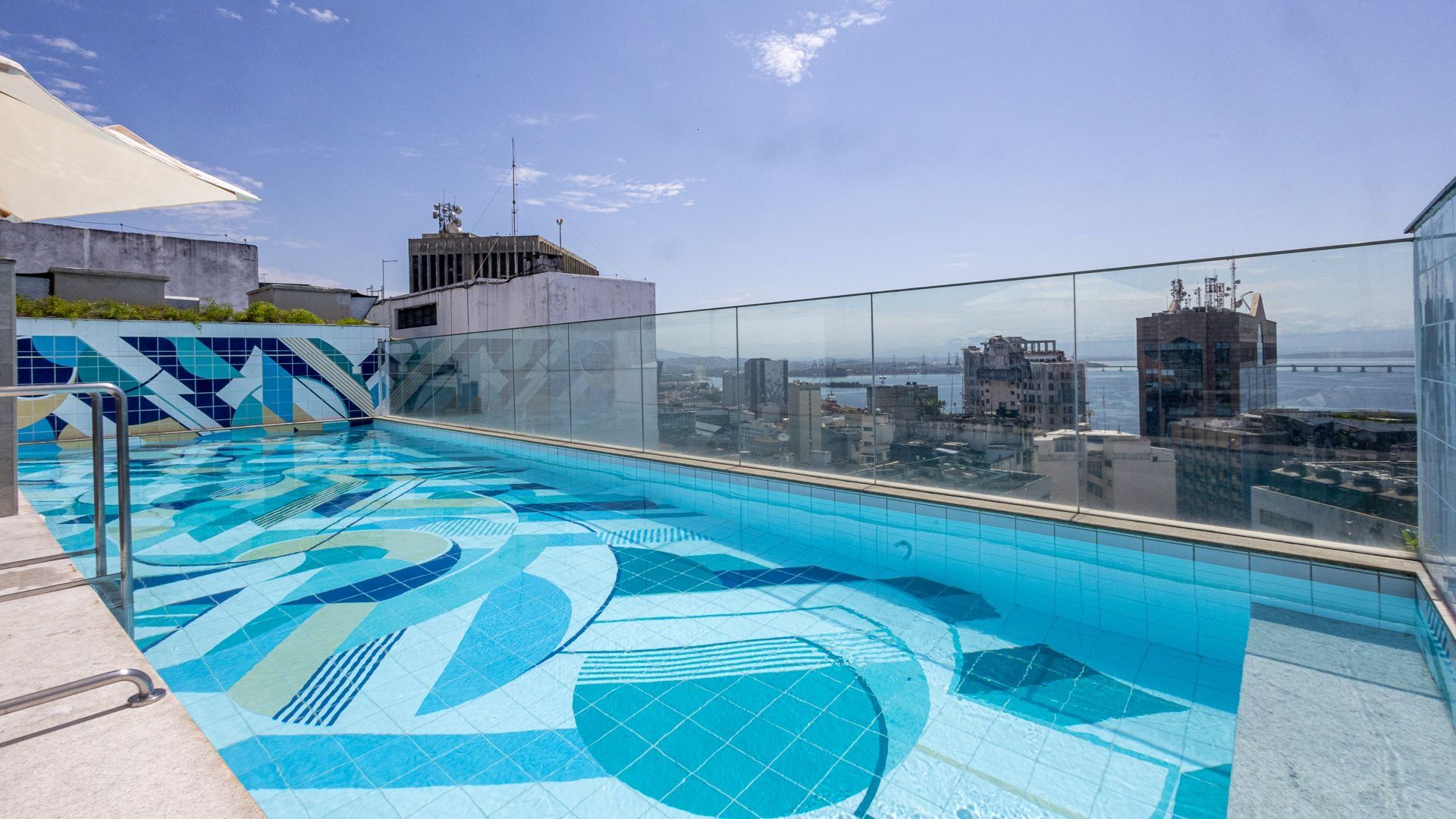 A rooftop swimming pool with a city view, glass railings, and a tiled floor.