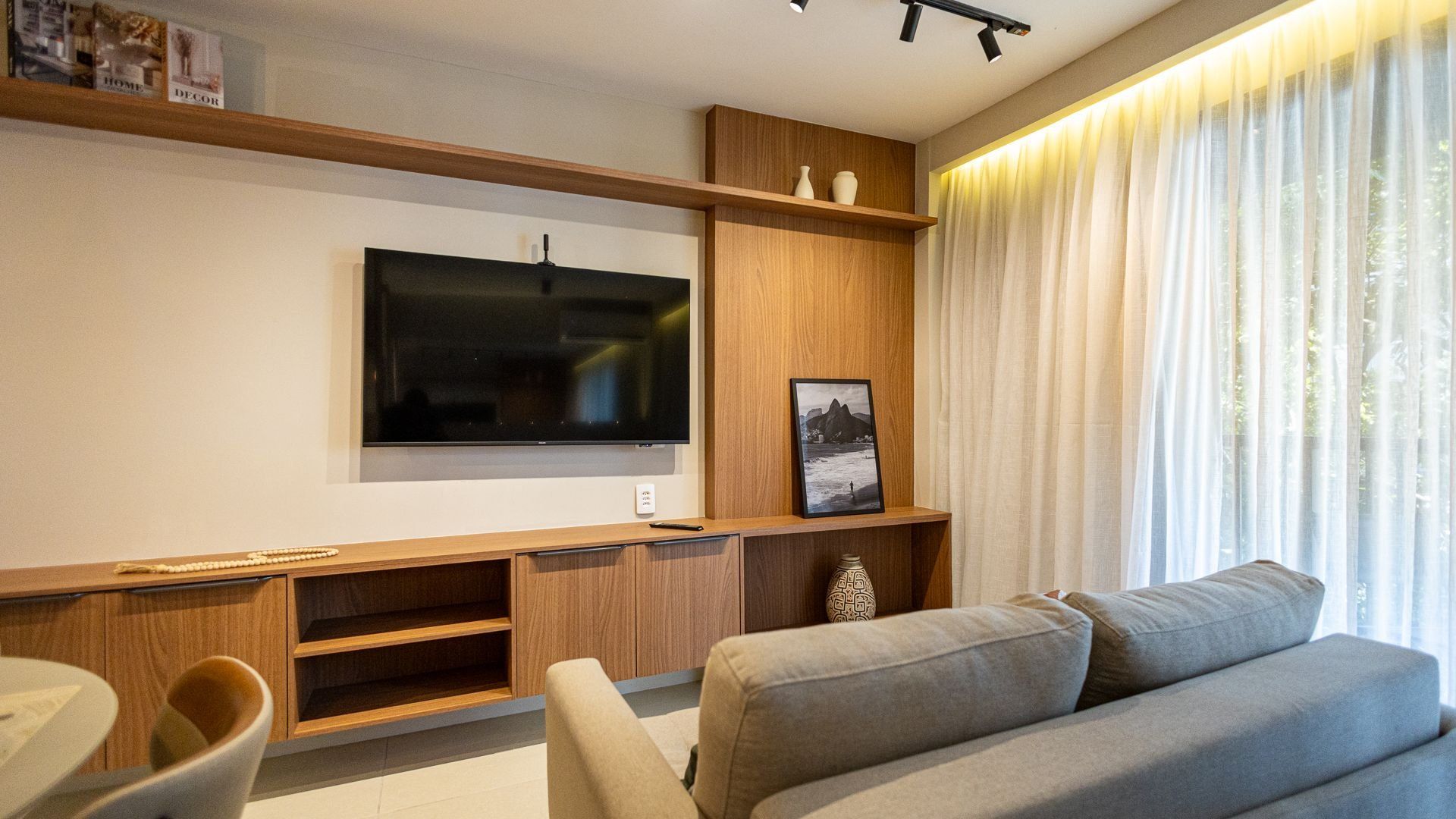 A modern living room with a flat-screen TV mounted on a wooden wall, a grey sofa, and a large window with sheer curtains.