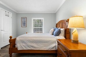 A bedroom with a bed, a bedside lamp, a dresser, a window with blinds, and a door. - Fin & Feather - Steinhatchee Florida (Steinhatchee)