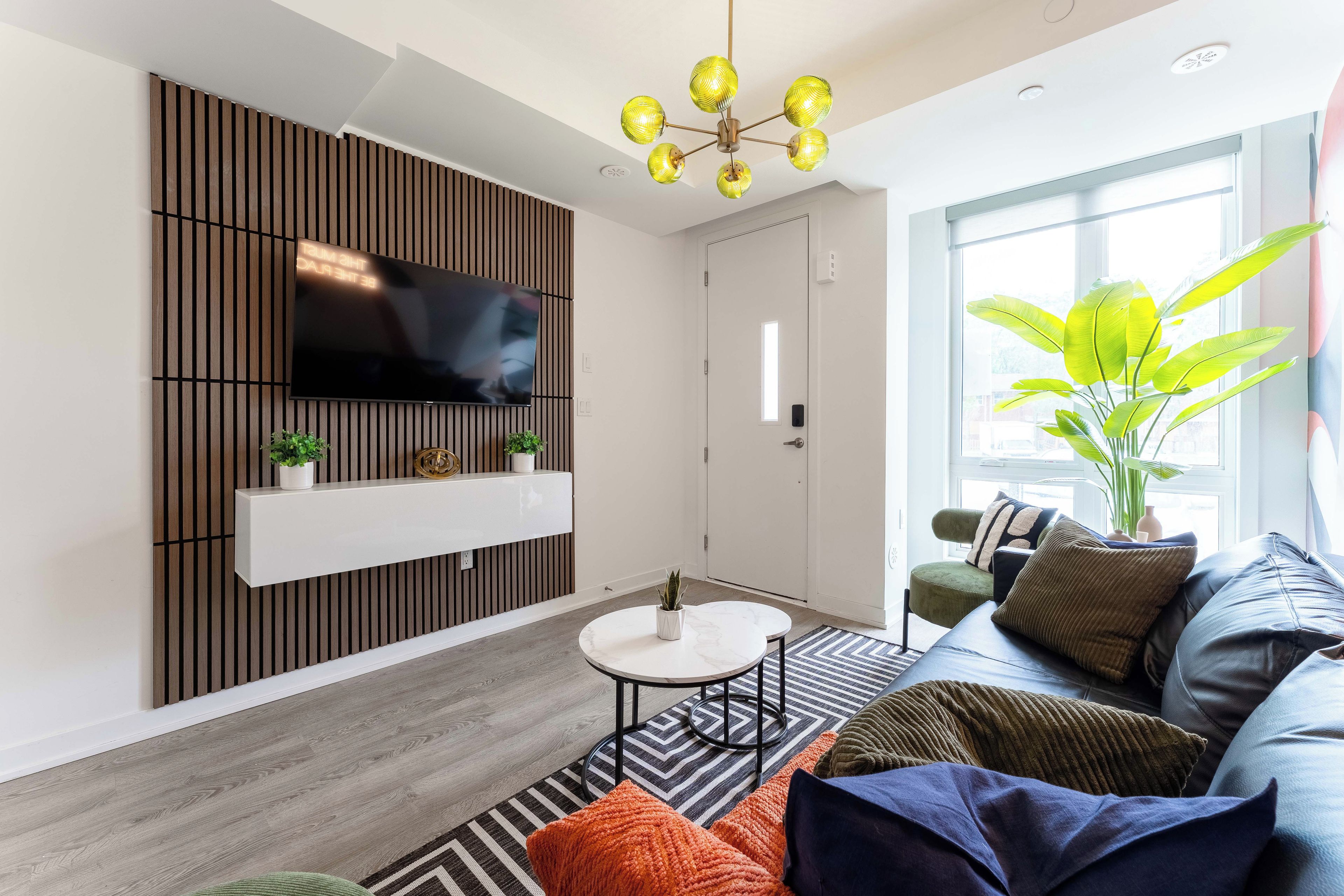 Ein modernes Wohnzimmer mit Flachbildfernseher an einer Wand aus Holzpaneelen, einem runden Couchtisch, einer Couch mit verschiedenen Kissen und einem großen Fenster mit Aussicht.
