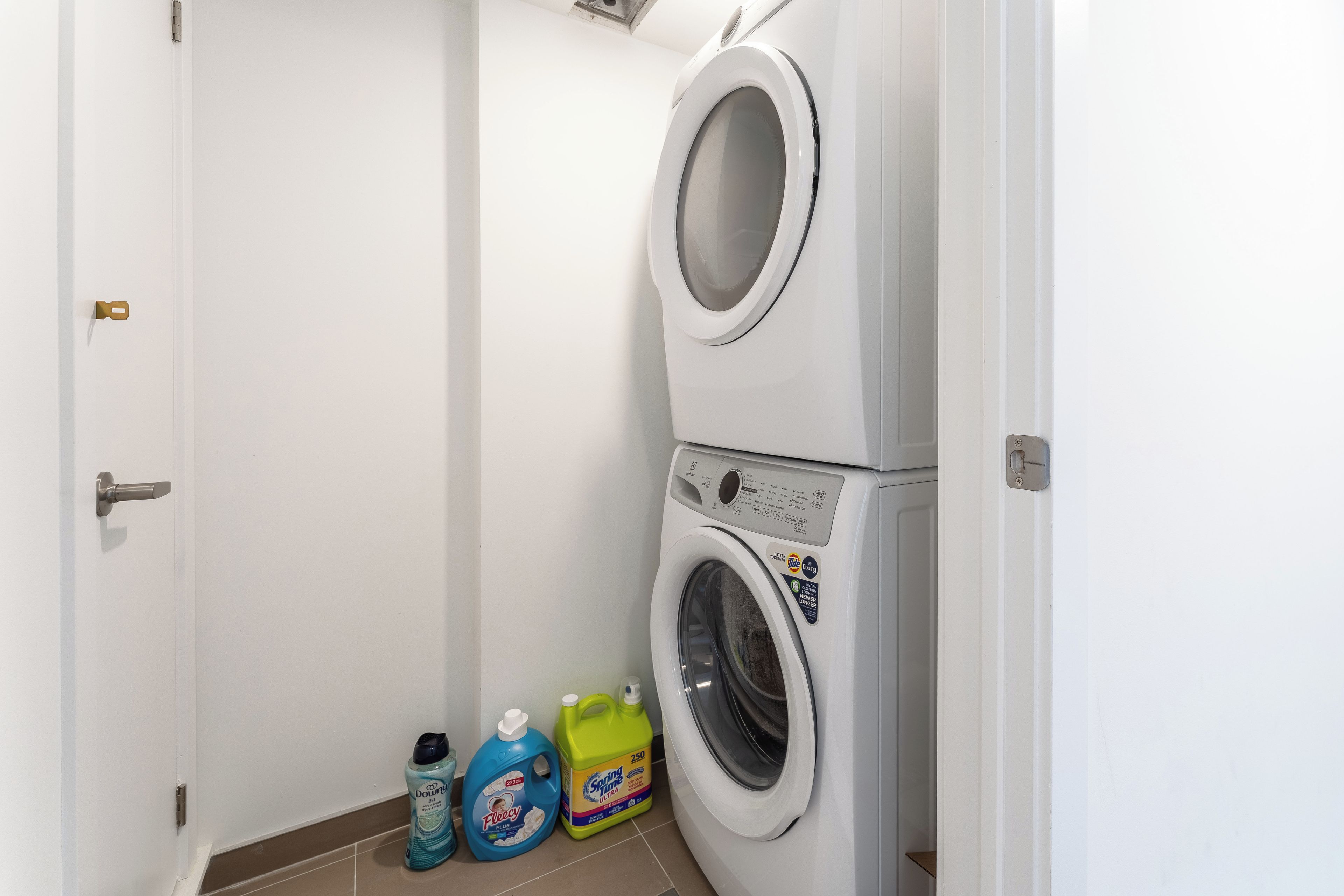 A laundry room with a front-loading washer and dryer, a door, and two bottles of laundry detergent.
