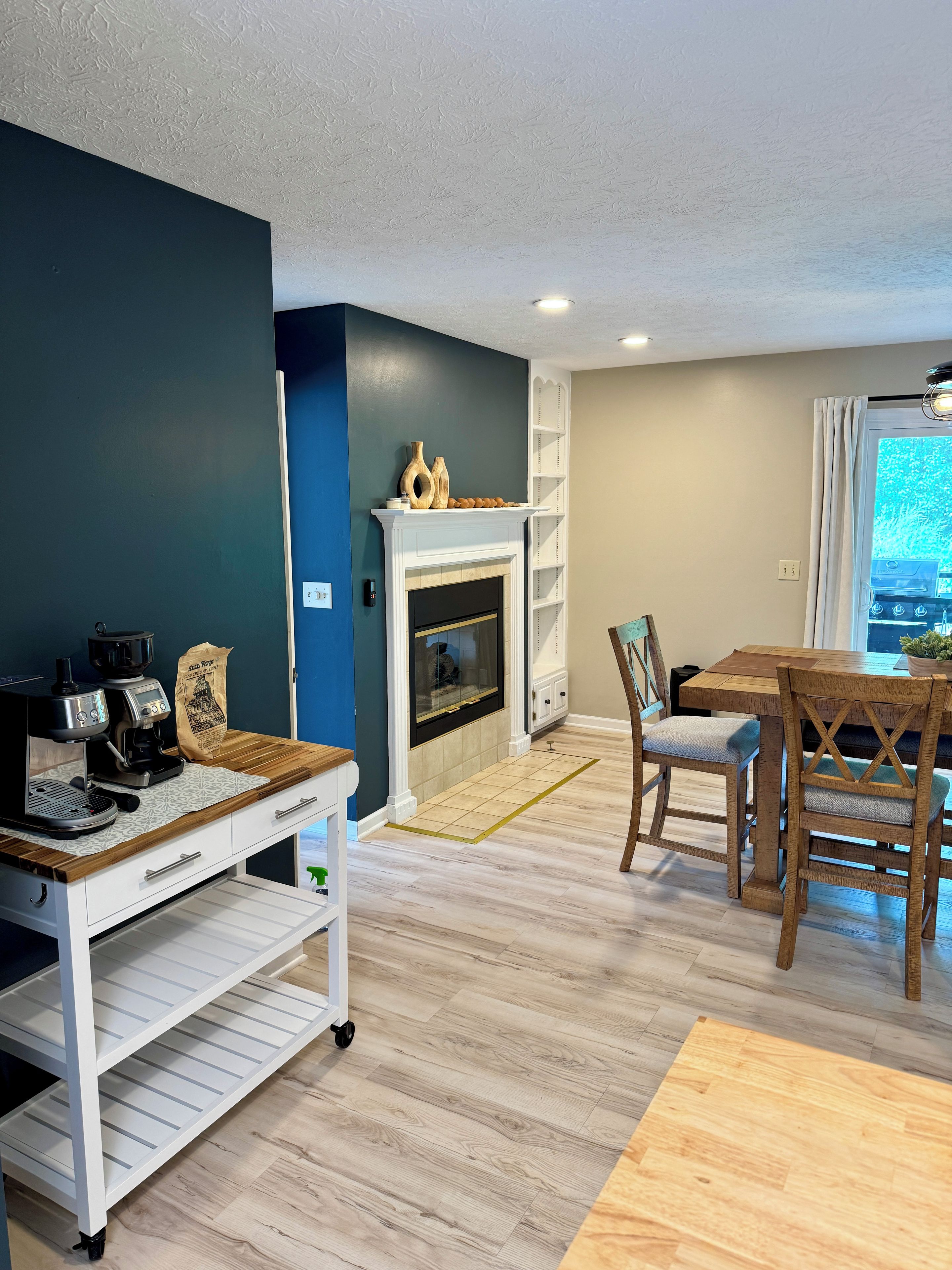 A modern living room with a fireplace, a coffee station, and a dining area with wooden chairs and a table.