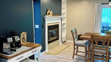 A modern living room with a fireplace, a coffee station, and a dining area with wooden chairs and a table.