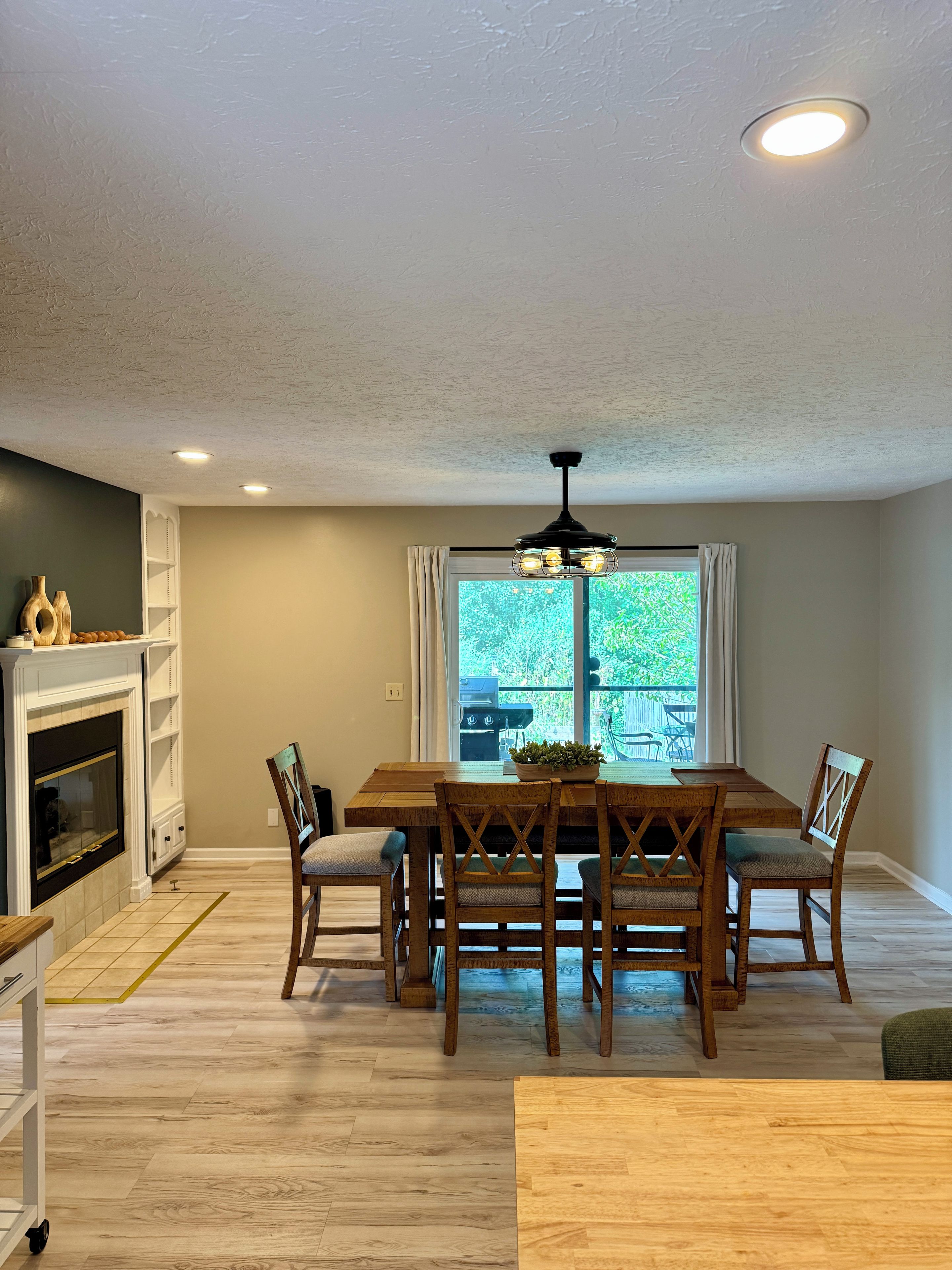 A dining area with a wooden table and chairs, a ceiling fan, and a view of the outdoors through a window.