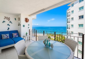 Een balkon met uitzicht op de oceaan, een ronde glazen tafel, rieten stoelen, een blauwe bank en decoratieve wandkunst.