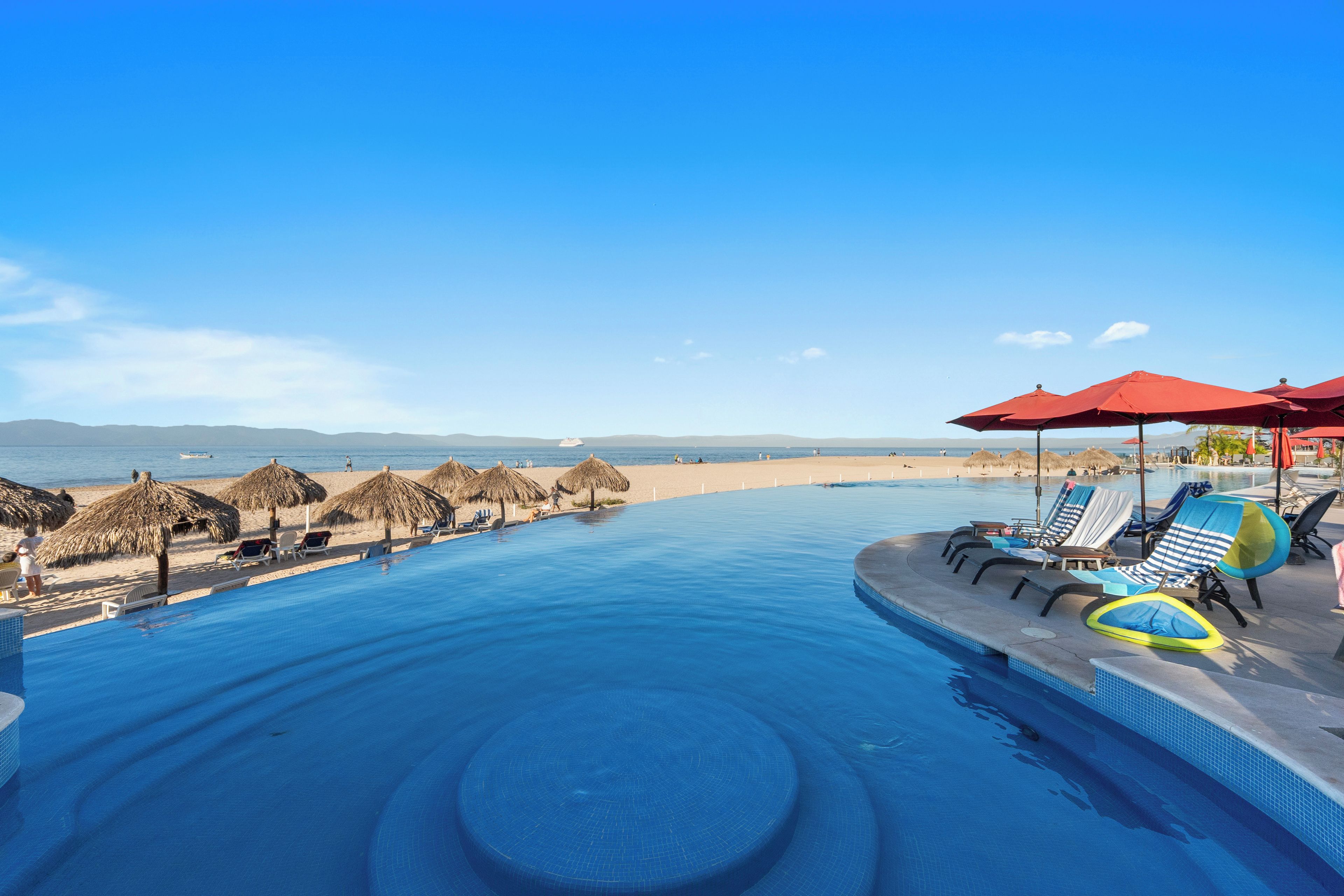 Ein Schwimmbecken mit Liegestühlen und Sonnenschirmen, ein Strandabschnitt mit Reetdachschirmen und ein freier Blick auf das Meer und ferne Berge.