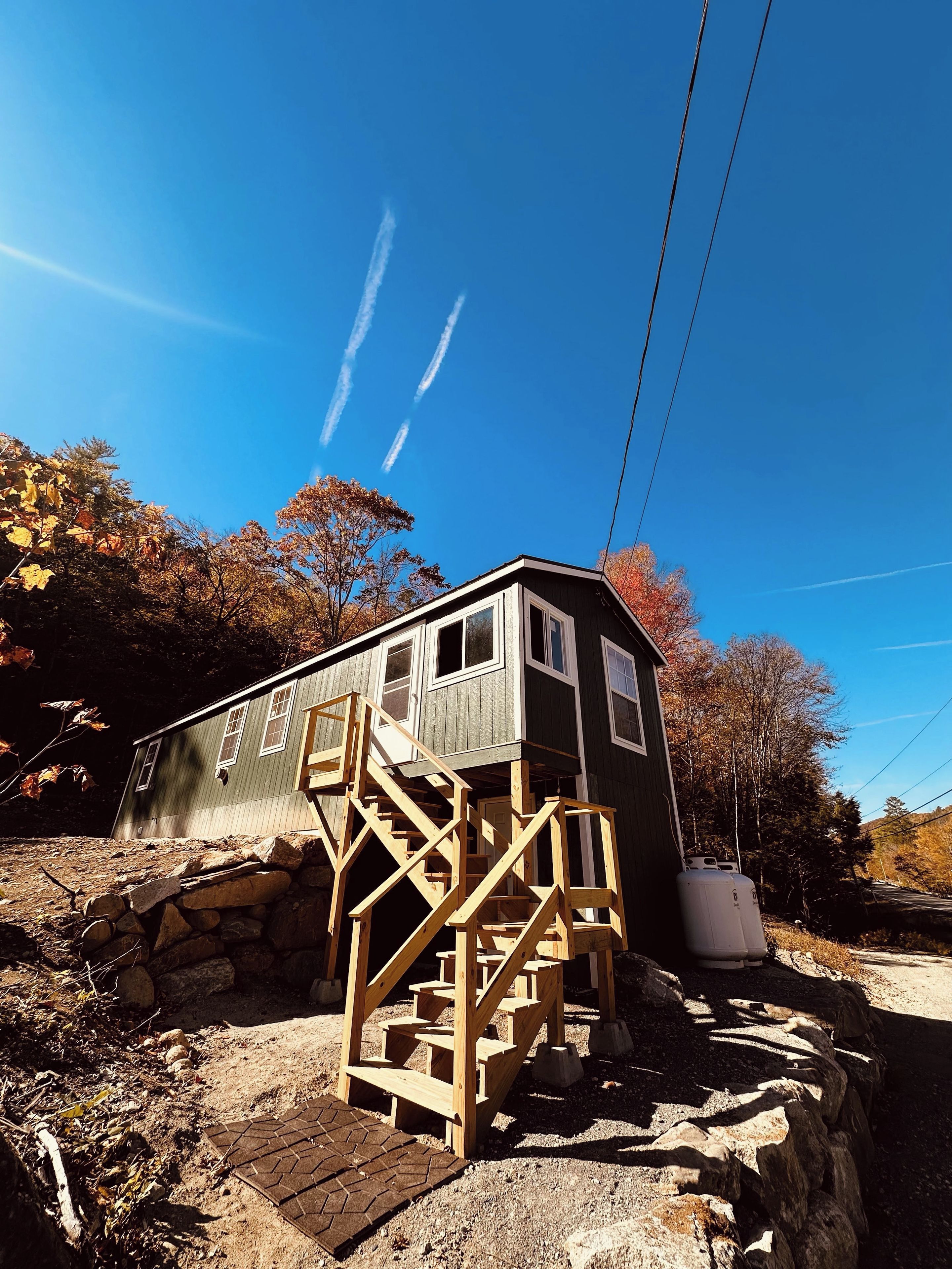 A cabin with a wooden staircase leading to a deck, surrounded by autumn foliage and a clear sky.