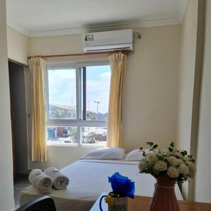 A hotel room with a bed, a window with curtains, an air conditioning unit, and a vase with flowers.