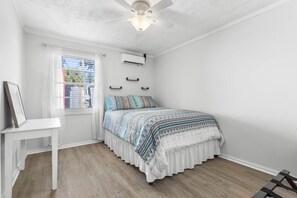 A bedroom with a bed, a ceiling fan, an air conditioning unit, a window with blinds, and a white table. - The Red Roof River Cottage - Home in Steinhatchee (Steinhatchee)