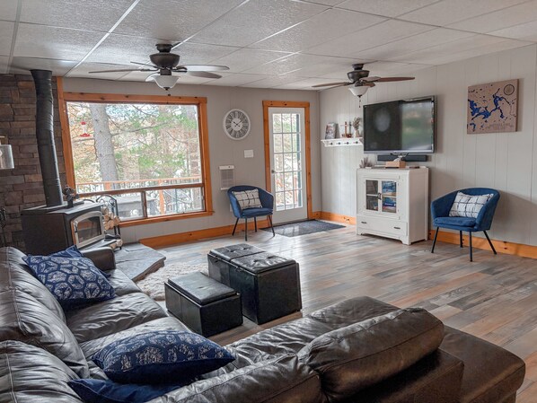 A living room with a fireplace, leather sofa, ottomans, a television, and a large window. - Baptiste Lake House In Bancroft- Waterfront- Pet Friendly (Hastings Highlands)