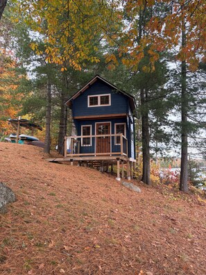 A cabin with a wooden deck, surrounded by trees and a hill. - Baptiste Lake House In Bancroft- Waterfront- Pet Friendly (Hastings Highlands)
