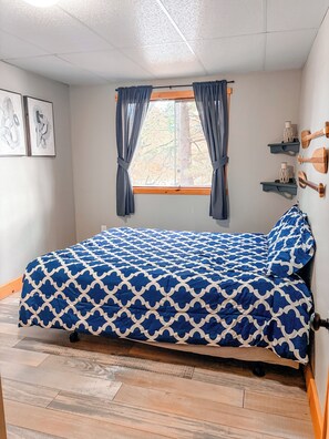 A room with a bed, a window with curtains, a wall-mounted shelf with decorative items, and a framed picture. - Baptiste Lake House In Bancroft- Waterfront- Pet Friendly (Hastings Highlands)