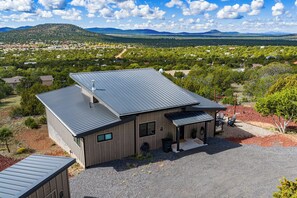A modern two-story house with a metal roof, surrounded by a gravel driveway and a landscaped area with trees and shrubs. - NEW! Hillside Home w/Hot Tub, Mountain Views, EV Charging (Williams)