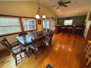 A dining area with a large wooden table and chairs, a ceiling fan, and a kitchen area with bar stools. - Cozy Ski Mountain House (Wardsboro)