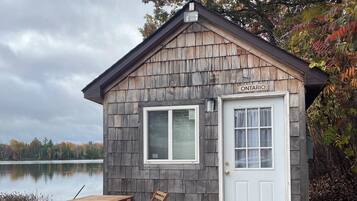 A small wooden cabin with a dock, a table, and chairs, located near a lake.