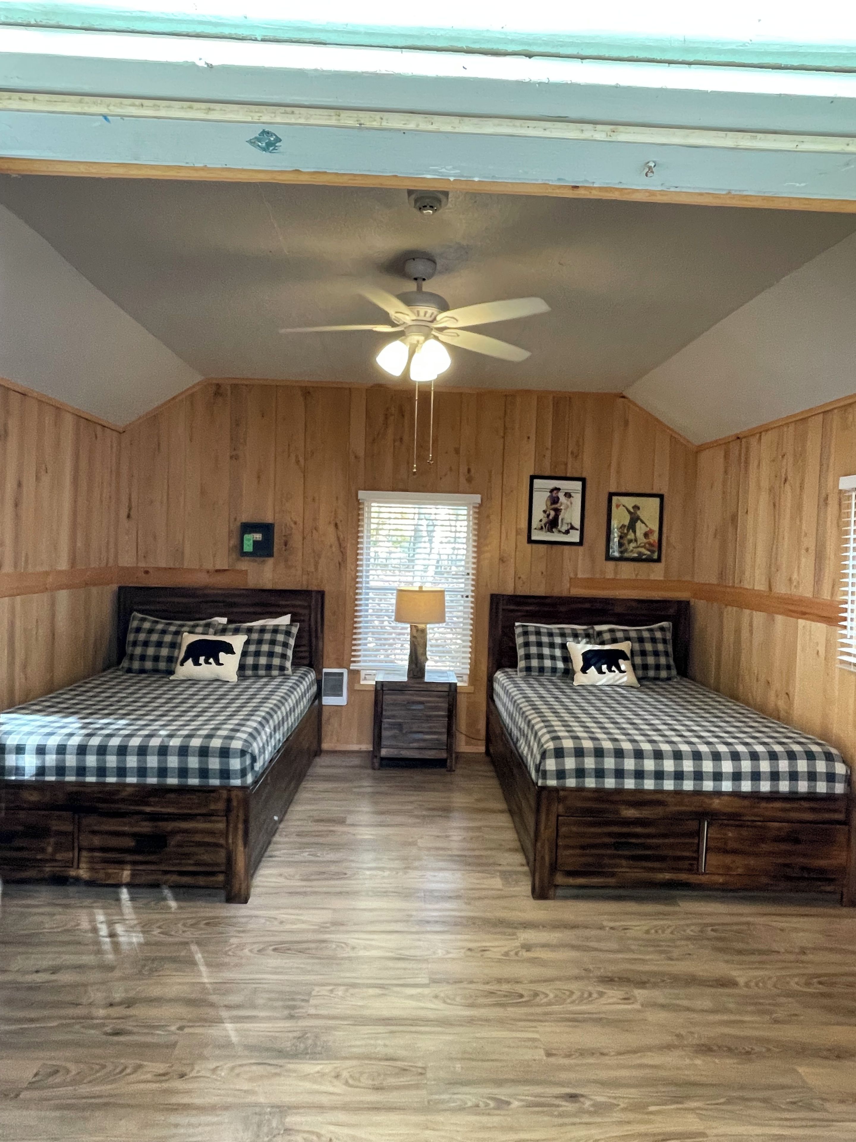 A room with two wooden beds, a ceiling fan, a window with blinds, and a wall-mounted light fixture.