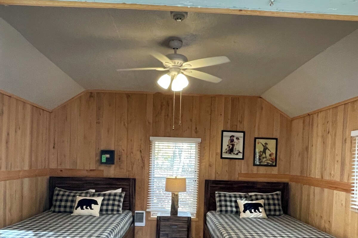 Una habitación con dos camas de madera, un ventilador de techo, una ventana con persianas y una lámpara fijada a la pared.