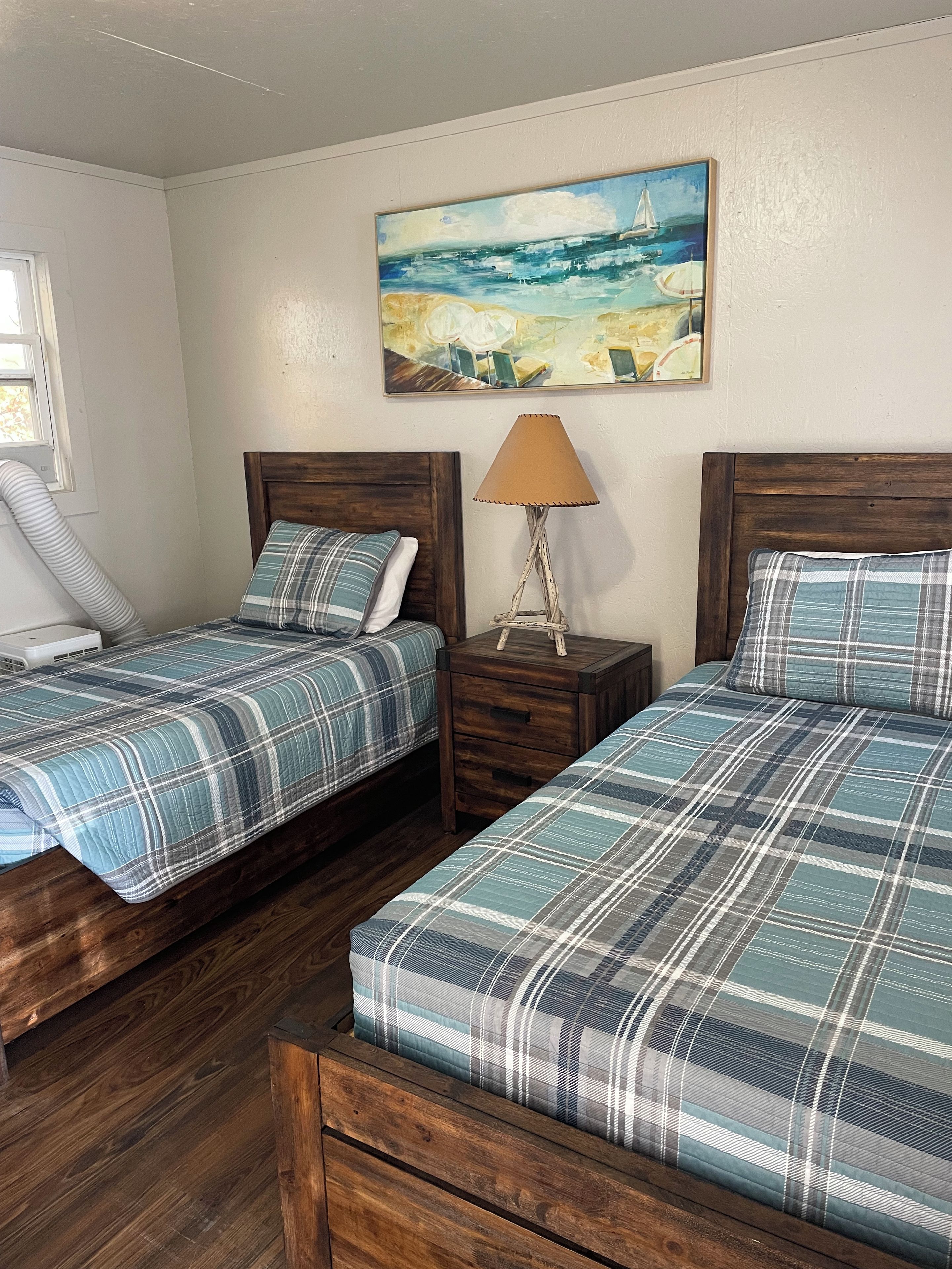 A bedroom with two beds, a nightstand with a lamp, a painting on the wall, and a window with a curtain.