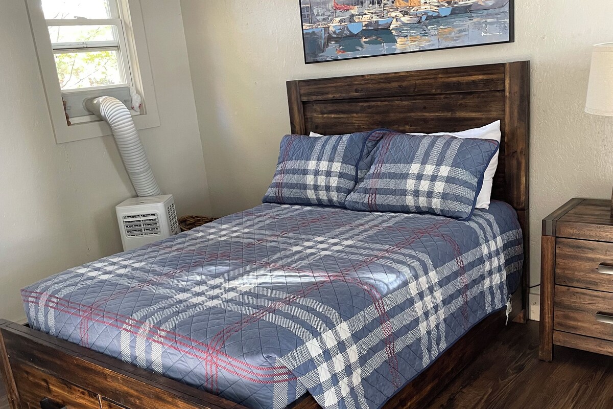Un dormitorio con cama de madera, ventana con vista a barcos, un cuadro en la pared y una mesita de noche.